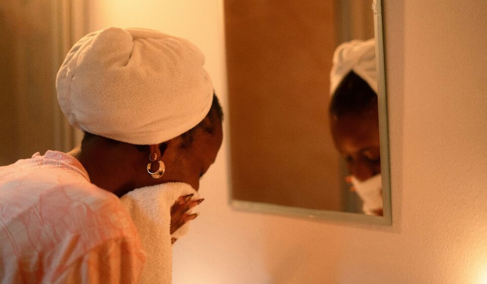 Woman with a towel wrapped around her hair drying her face in front of a mirror